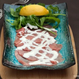 Thin slices of raw beef arranged on a blue plate with Cipriani sauce, rocket, and a lemon wedge.