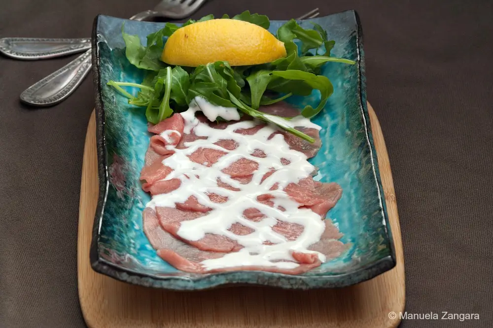 Thin slices of raw beef arranged on a blue plate with Cipriani sauce, rocket, and a lemon wedge.