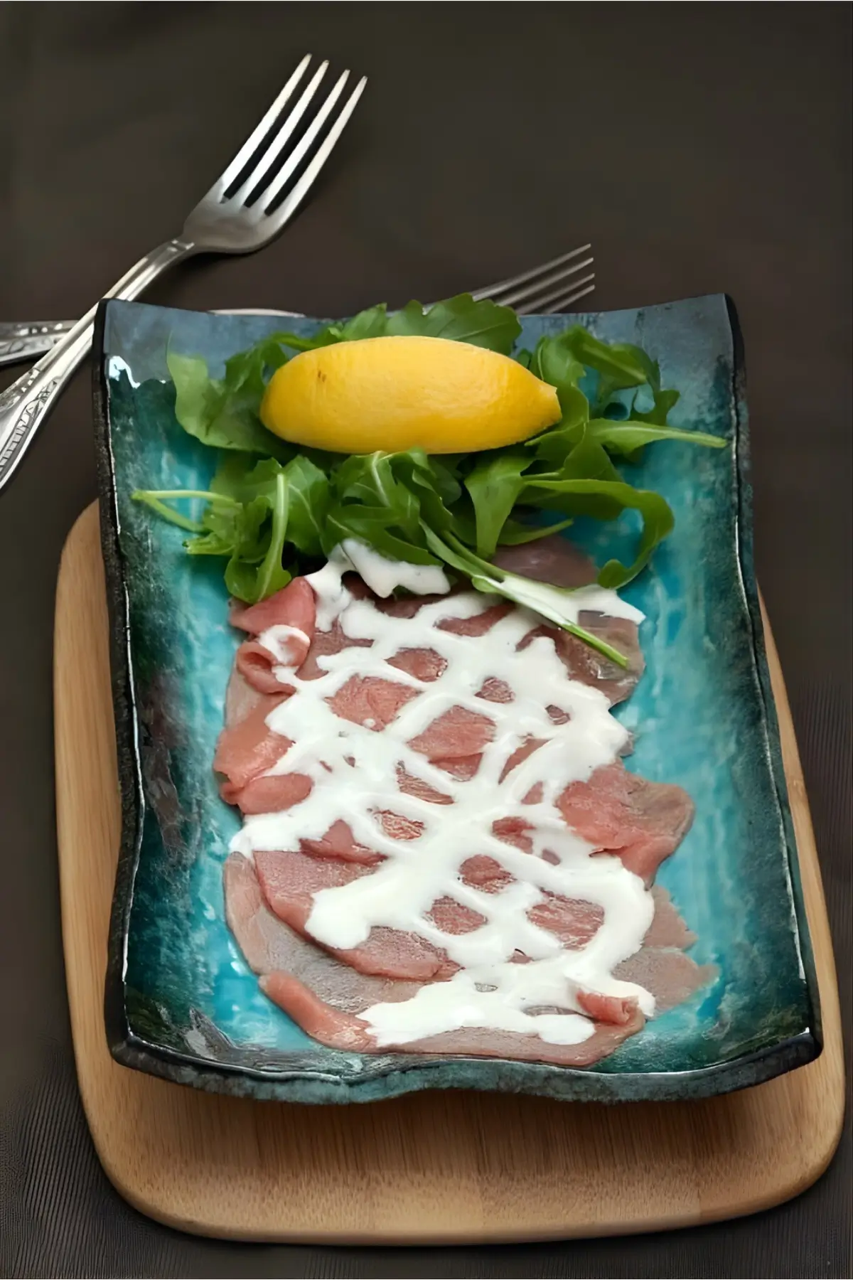 Plated Beef Carpaccio alla Cipriani, fresh rocket, and a lemon wedge on a wooden board.