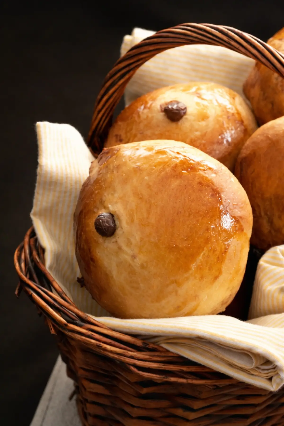 Golden chocolate chip buns with a shiny glaze in a woven basket lined with a cloth.