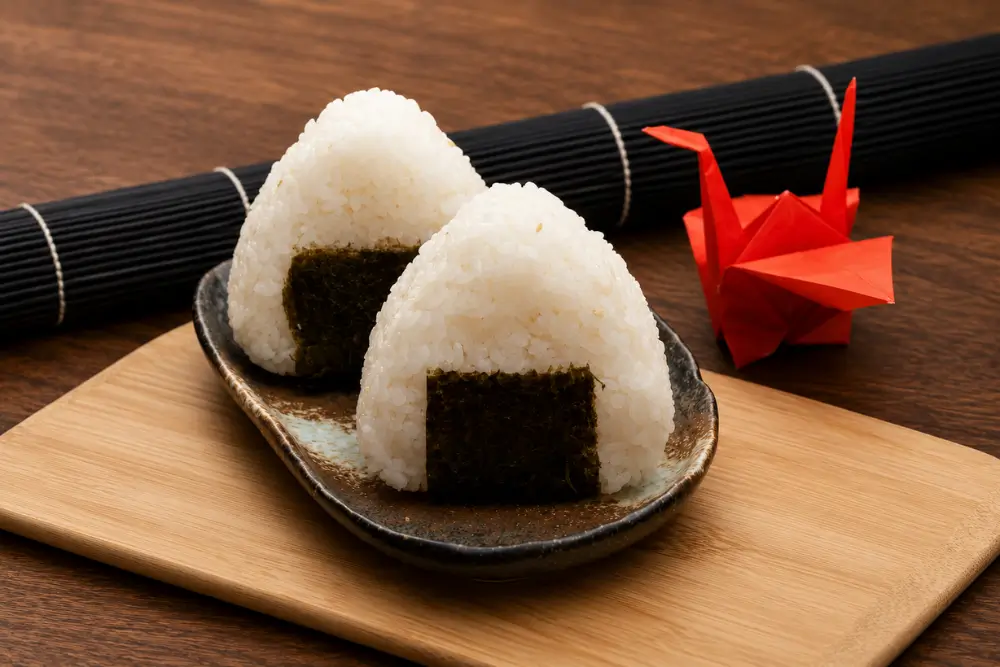 Two triangular rice balls with nori served on a ceramic plate with a red origami crane.