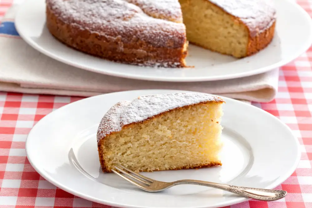 Slice of moist cake dusted with icing sugar served on a white plate with a fork.