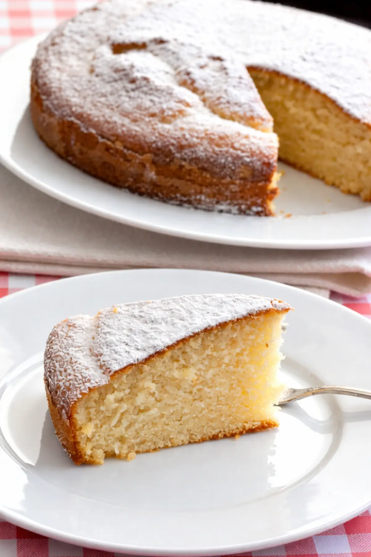 Slice of soft vanilla cake dusted with icing sugar served on a white plate.