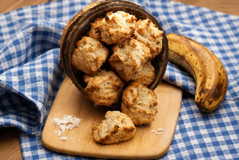 Golden brown banana coconut treats displayed in a coconut shell with a ripe banana in the background.