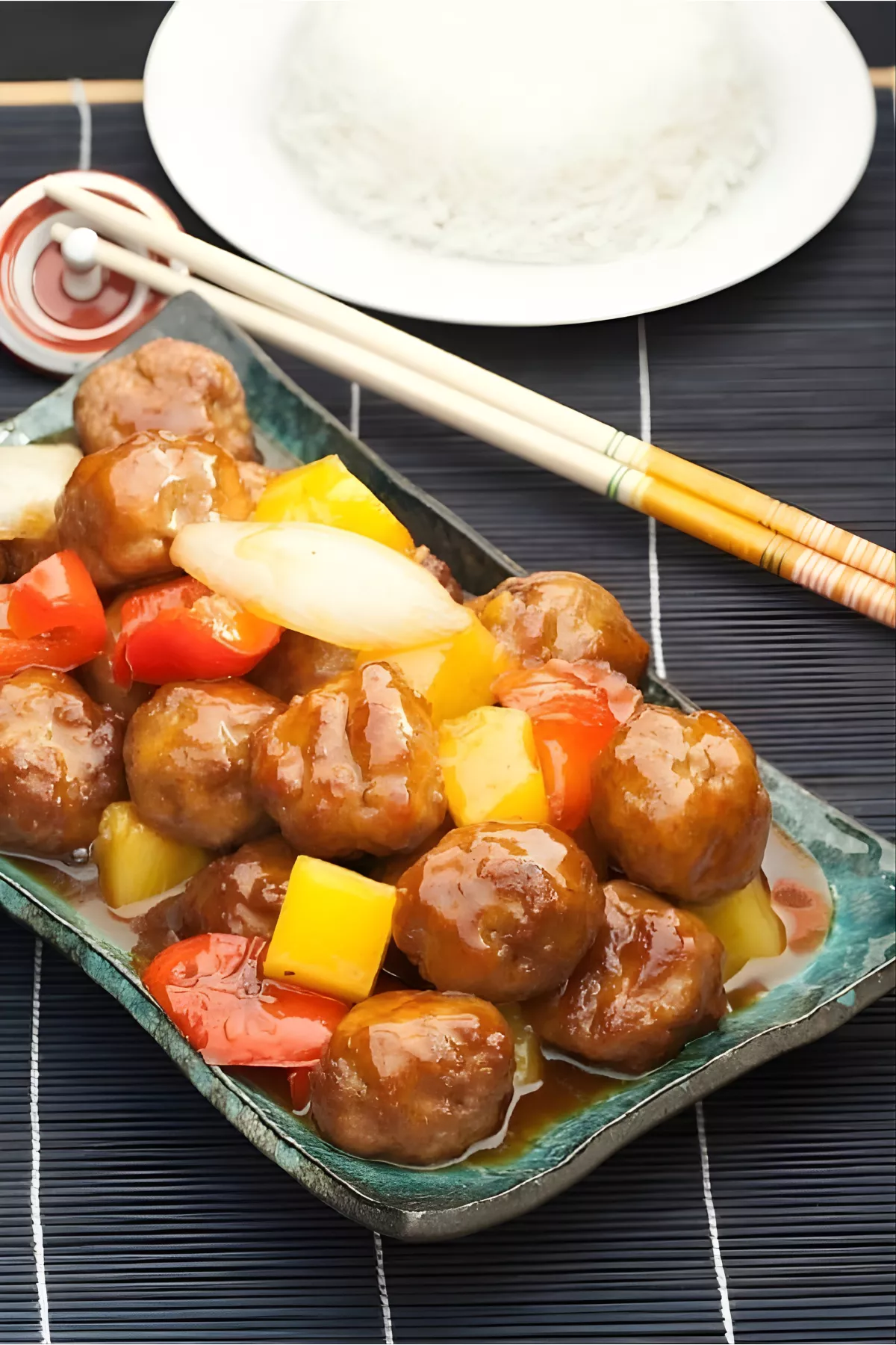 A close-up of sweet and sour pork meatballs served in a dish with chopsticks and a plate of rice in the background.