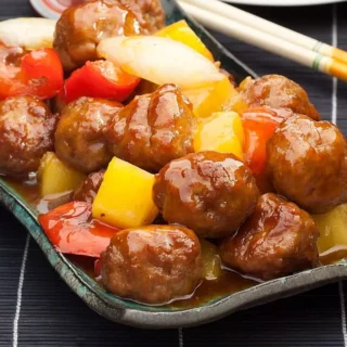 A close-up of Chinese sweet and sour pork meatballs with red peppers, pineapple, and onions served in a dish.