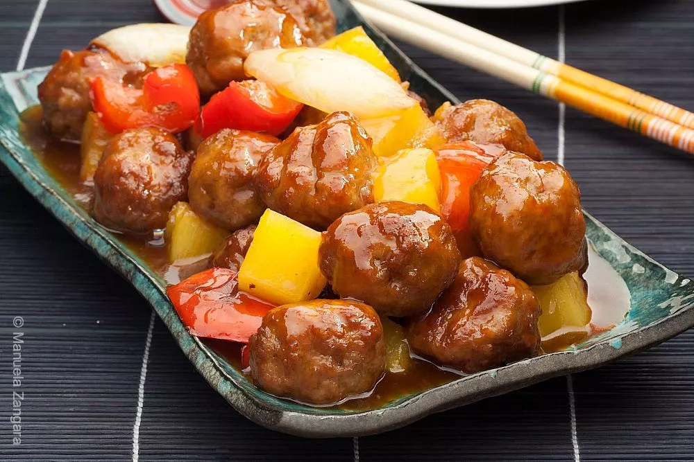 A close-up of Chinese sweet and sour pork meatballs with red peppers, pineapple, and onions served in a dish.