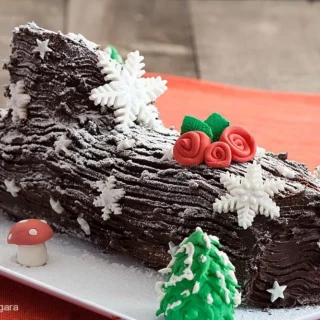 Close-up of the Bûche de Noël – rich chocolate ganache Yule Log Cake decorated with fondant snowflakes, red roses, and a small mushroom.