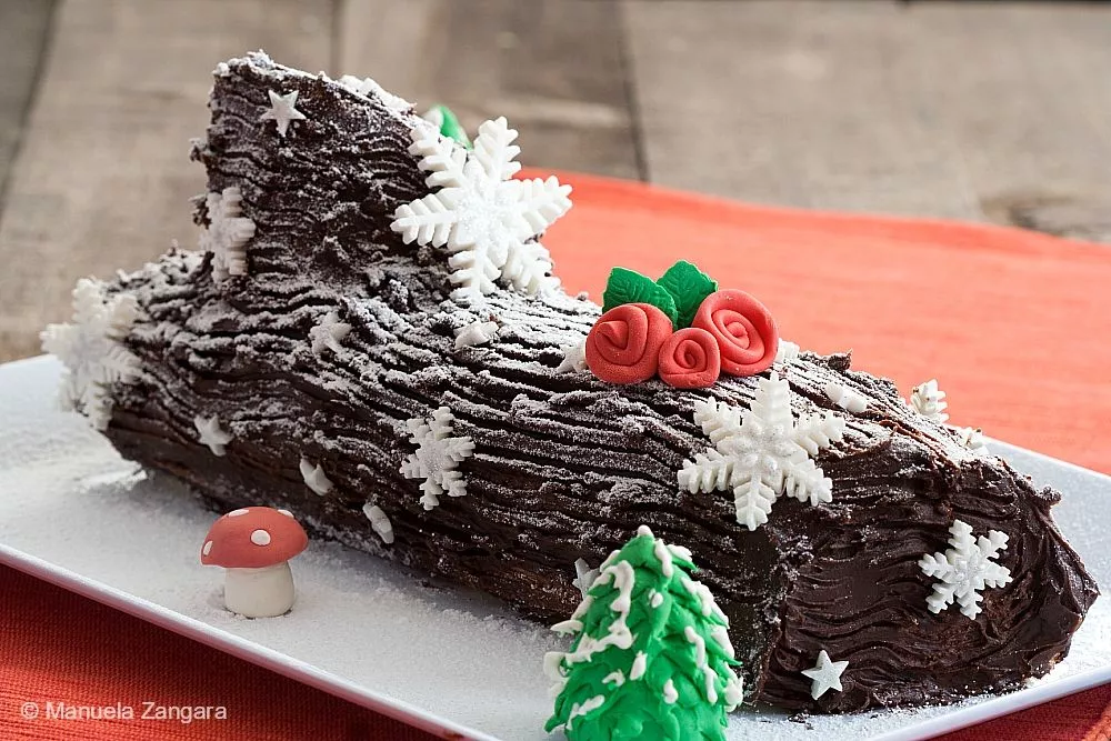 Close-up of the Bûche de Noël – rich chocolate ganache Yule Log Cake decorated with fondant snowflakes, red roses, and a small mushroom.