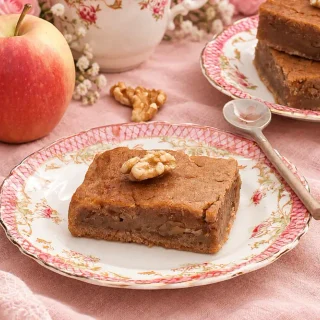 Vegan Apple Spice Cake topped with a walnut on a floral plate, served with a fresh apple and additional cake squares in the background.