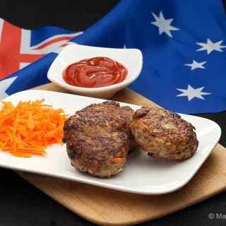 Juicy beef rissoles on a white plate with grated carrot and tomato sauce, served on a wooden board.
