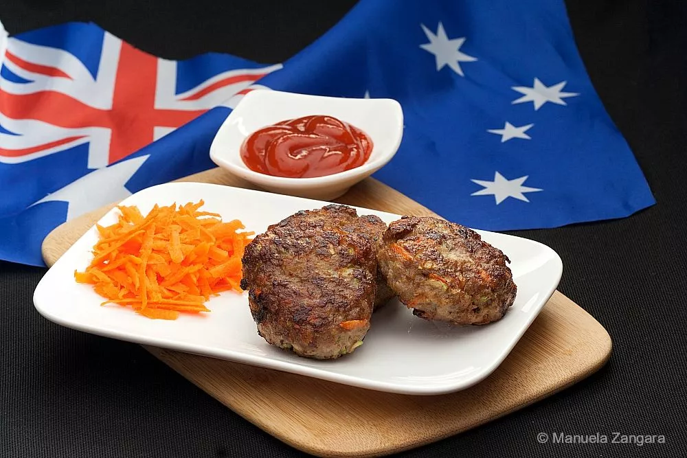 Juicy beef rissoles on a white plate with grated carrot and tomato sauce, served on a wooden board.