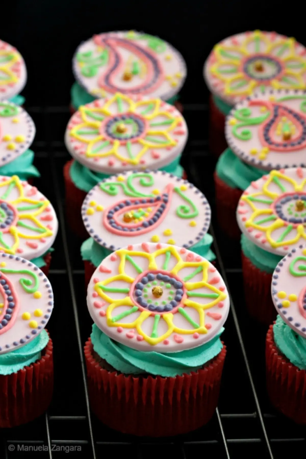 Close-up of a chocolate cupcake topped with teal buttercream and a fondant disc decorated with colourful henna-style piping.