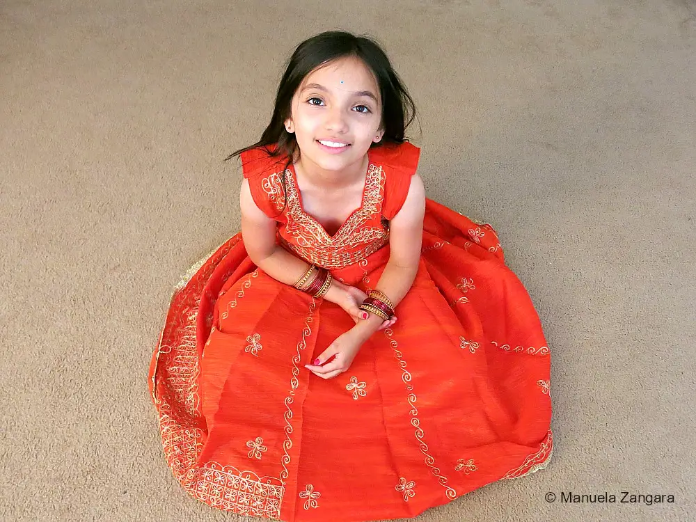 Girl dressed in an orange traditional Indian outfit sitting on the floor and smiling at the camera.