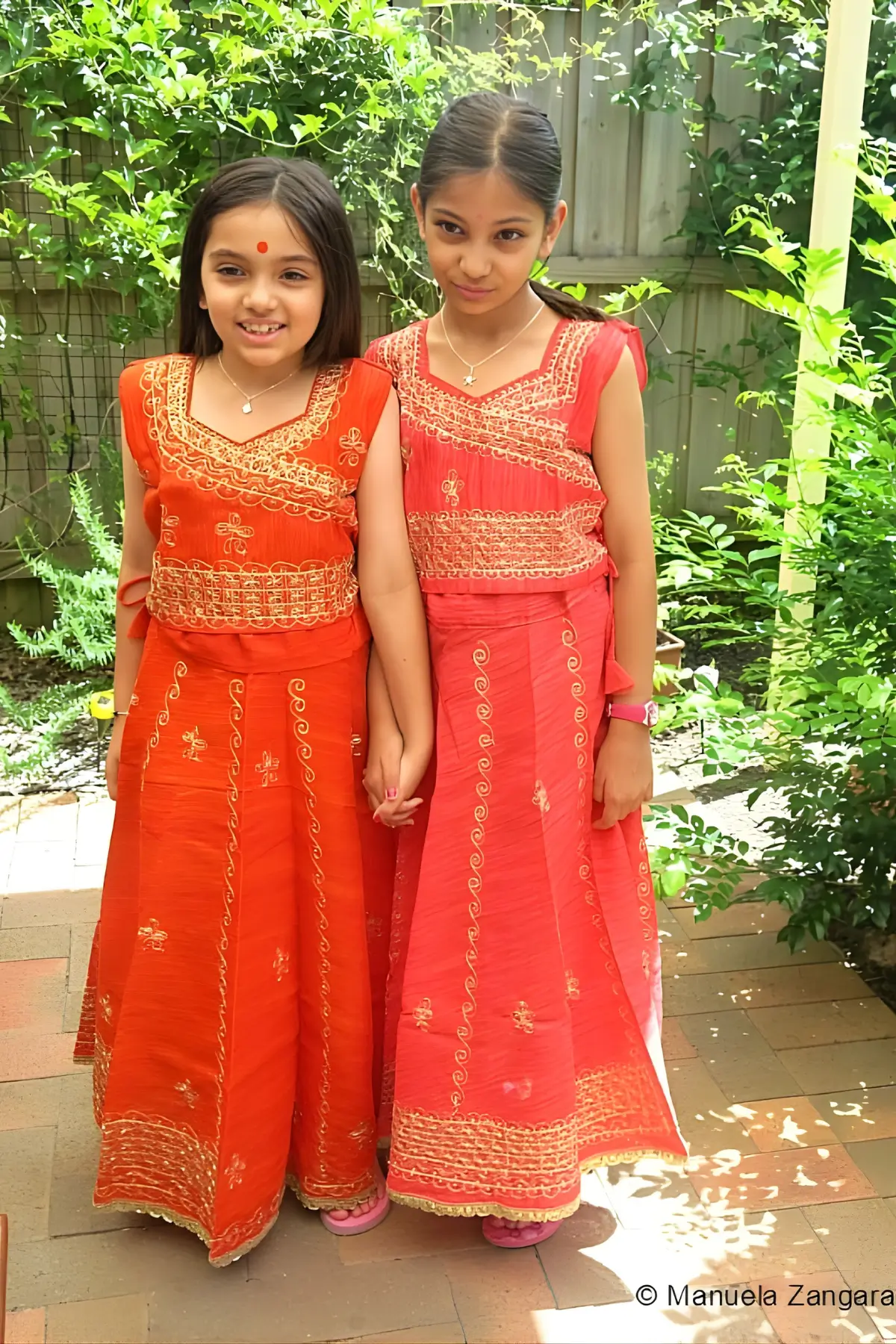 Two girls wearing bright orange and pink traditional Indian dresses, smiling and holding hands outdoors.