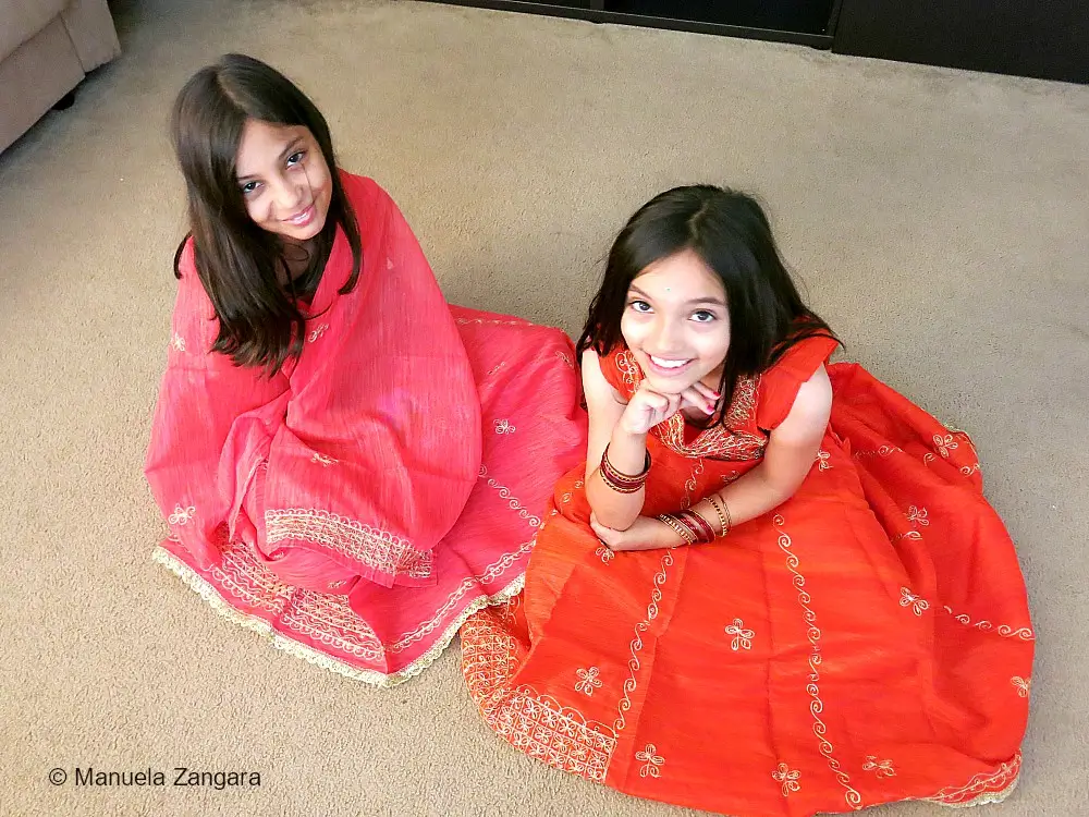 Two girls sitting on the floor wearing bright traditional Indian dresses with gold embroidery.