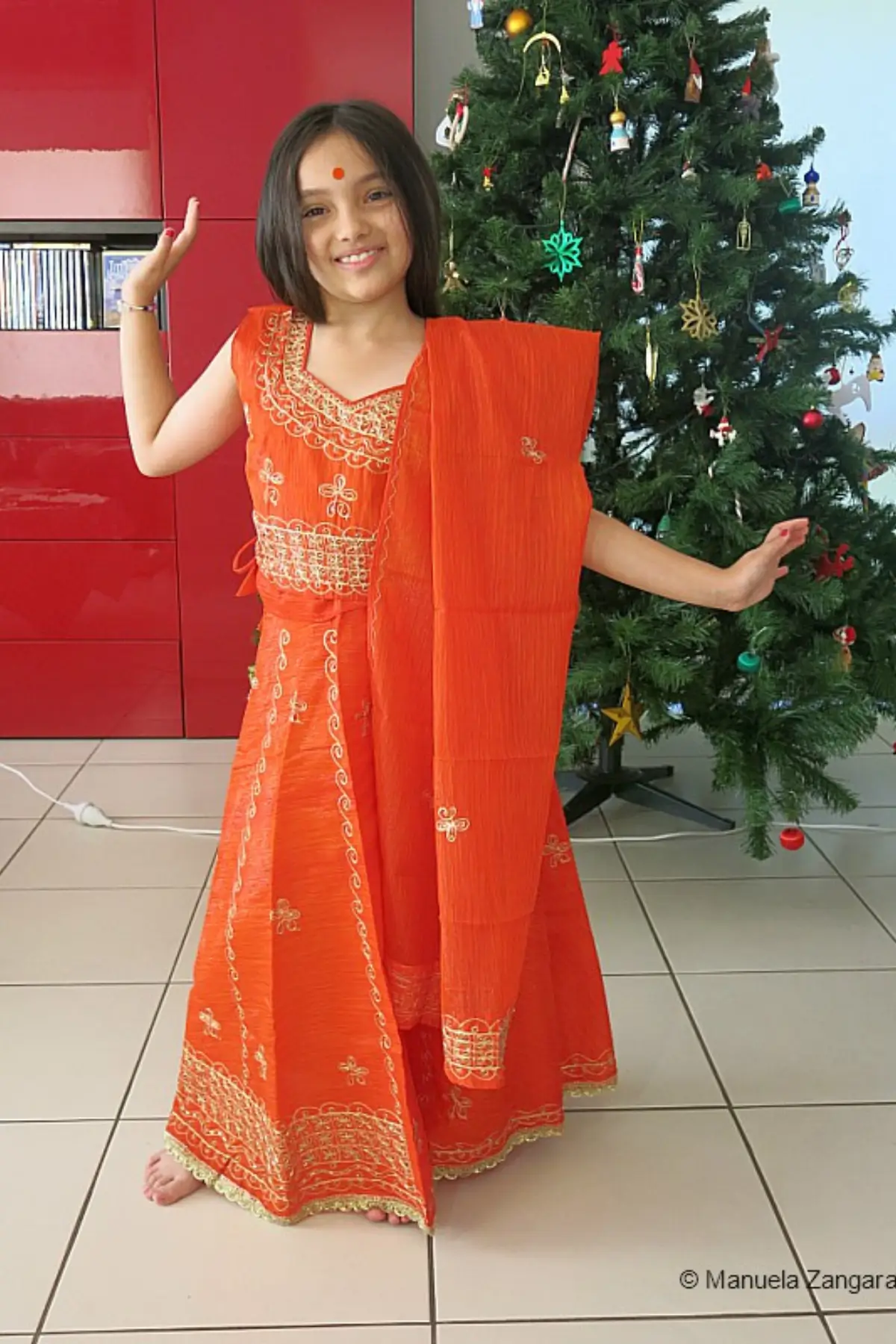 Young girl wearing a bright orange traditional Indian outfit standing beside a decorated Christmas tree.