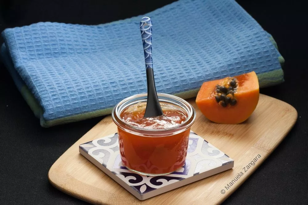 A jar of Papaya Jam with a spoon on a tiled coaster beside fresh papaya.