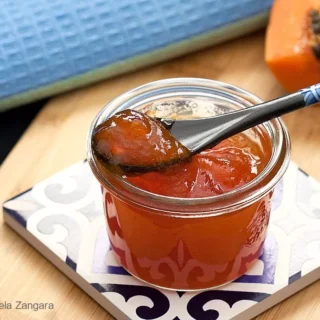 A glass jar of papaya jam placed on a decorative tile with a blue cloth in the background.