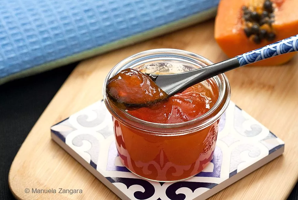 A glass jar of papaya jam placed on a decorative tile with a blue cloth in the background.