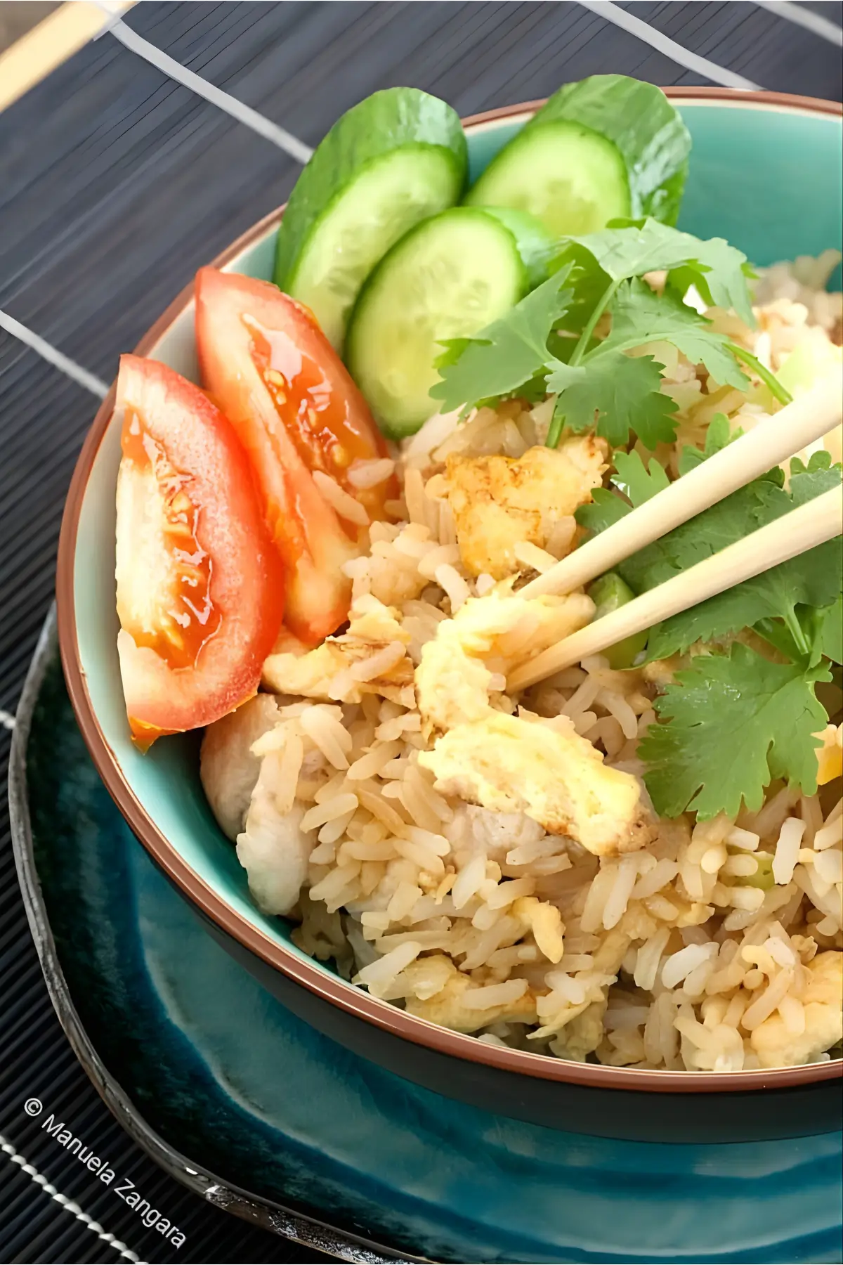Close-up of Thai chicken fried rice in a turquoise bowl, topped with coriander and served with tomato and cucumber slices.