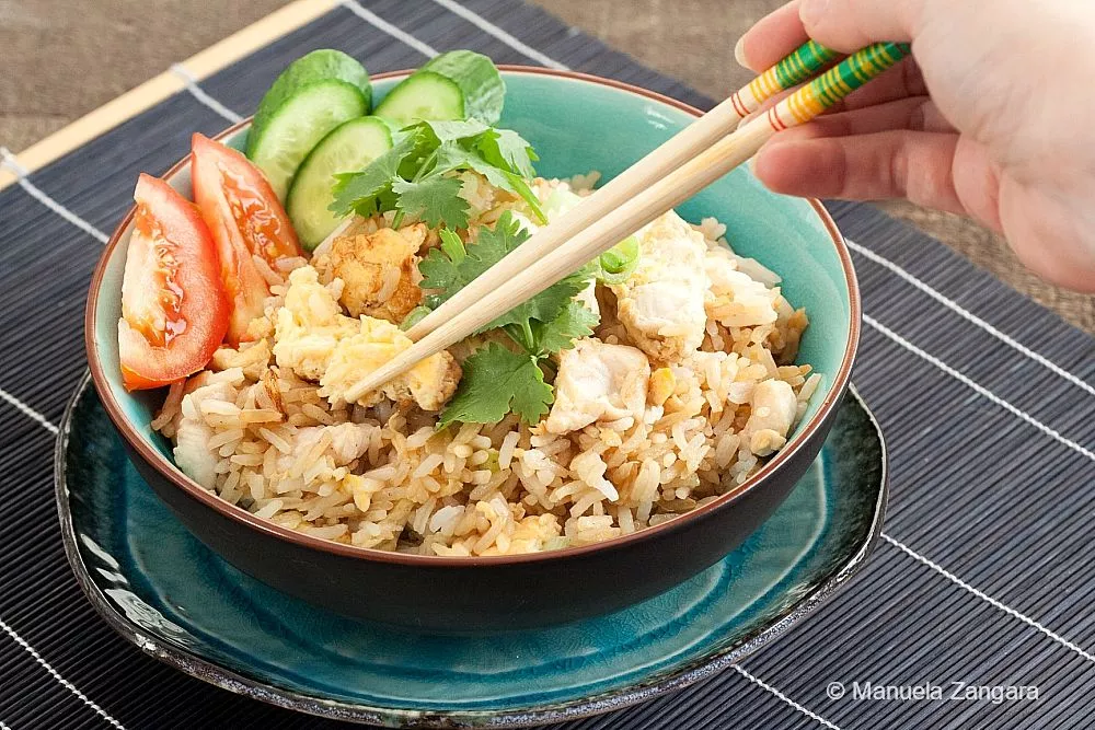 Hand picking up Thai chicken fried rice with chopsticks, served with cucumber, tomato, and coriander in a bowl.