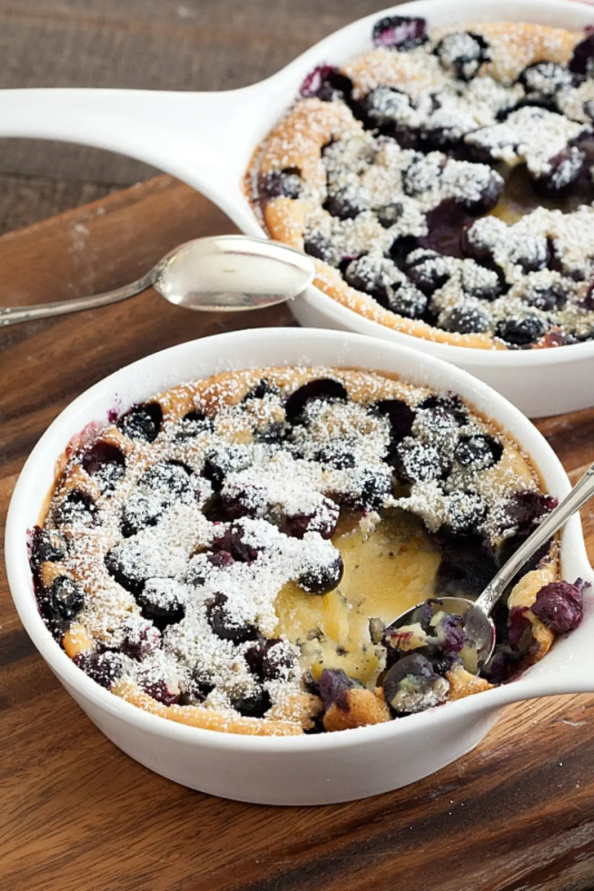 Blueberry Clafoutis baked in white ceramic dishes, dusted with icing sugar and served with a spoon.