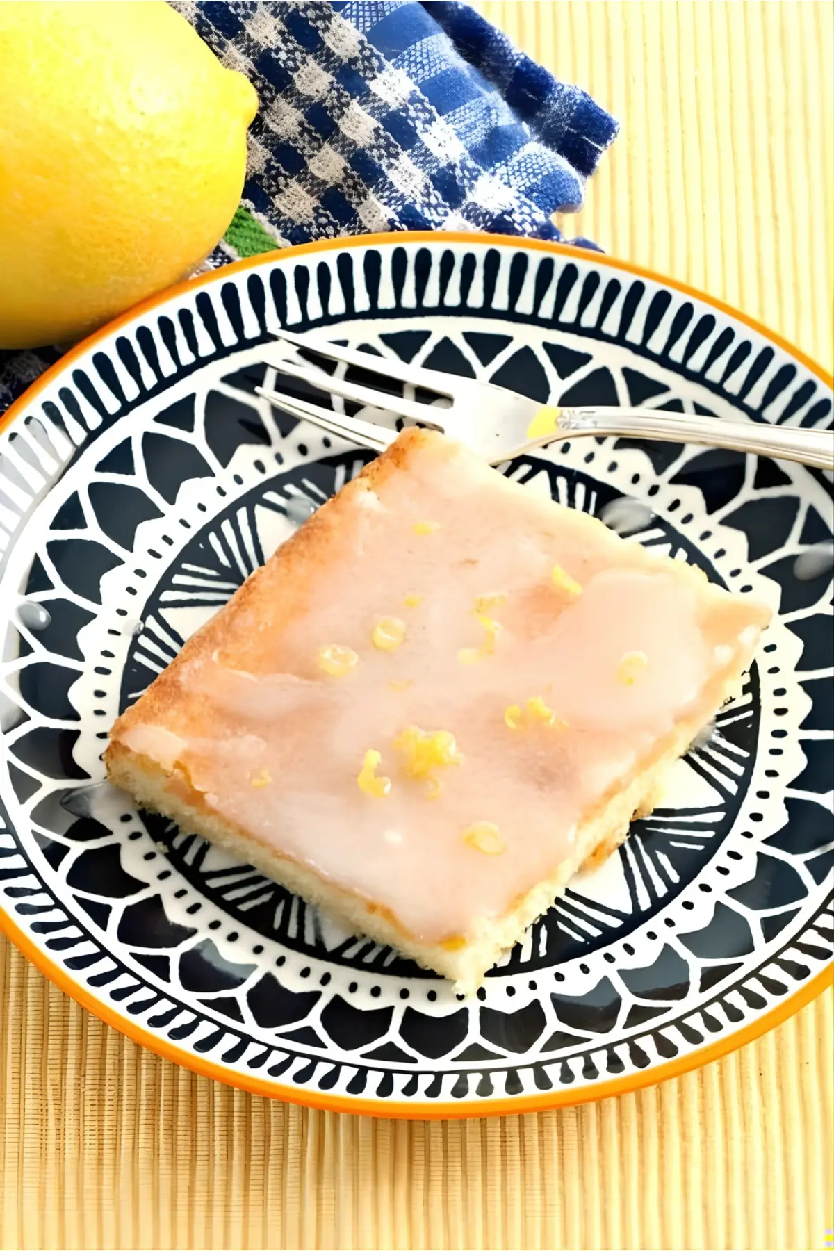 A piece of lemon cake with icing and lemon zest on a patterned plate.