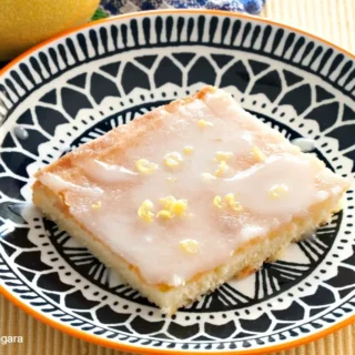 A glazed lemon cake square on a patterned plate with lemon zest on top.