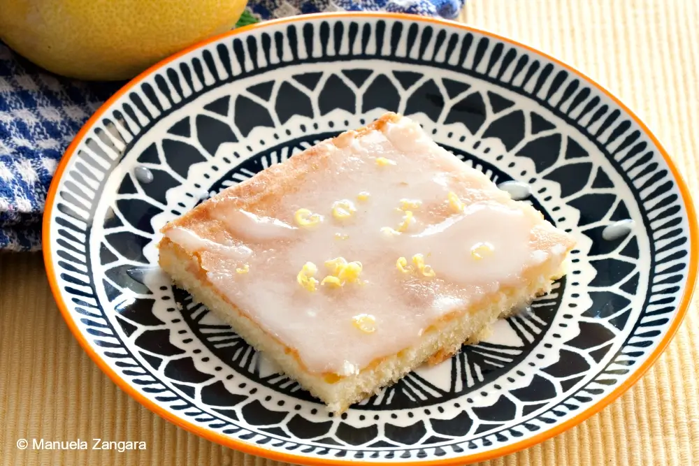 A glazed lemon cake square on a patterned plate with lemon zest on top.