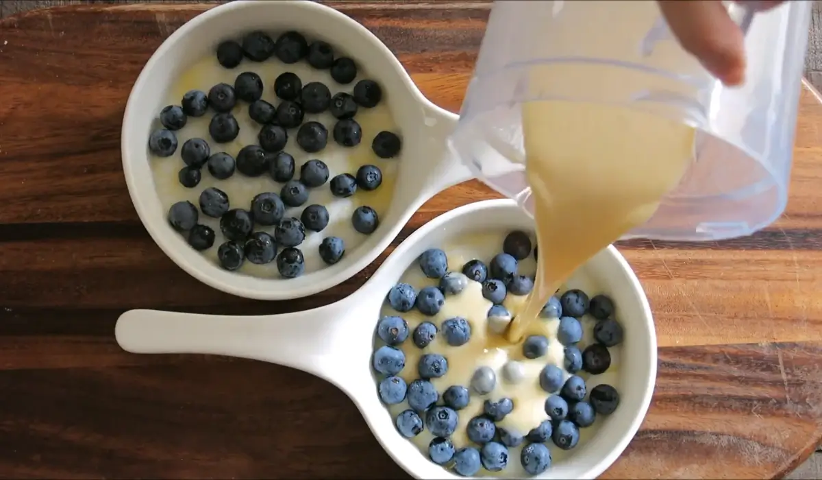 Batter being poured over fresh blueberries in white ceramic dishes.