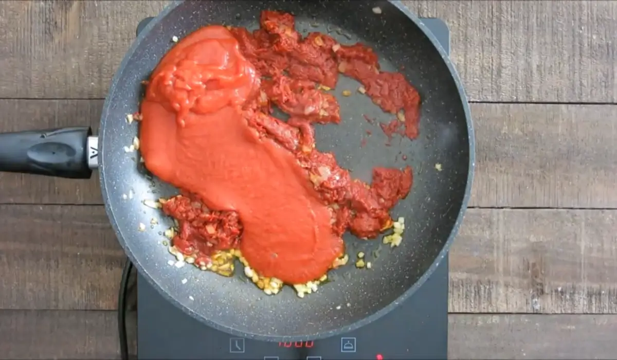 Tomato paste and tomato purée being added to sautéed onions in a pan.