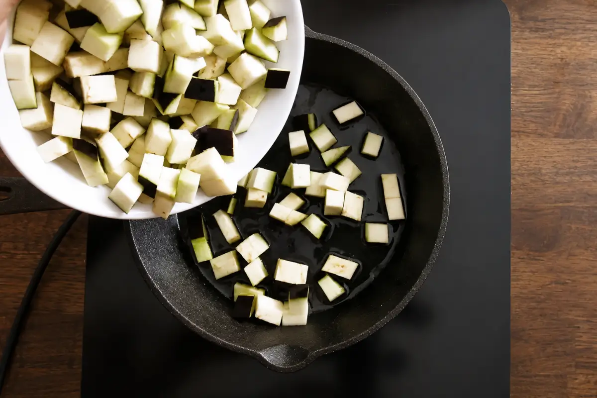 Diced eggplant being added to hot olive oil in a cast-iron skillet.