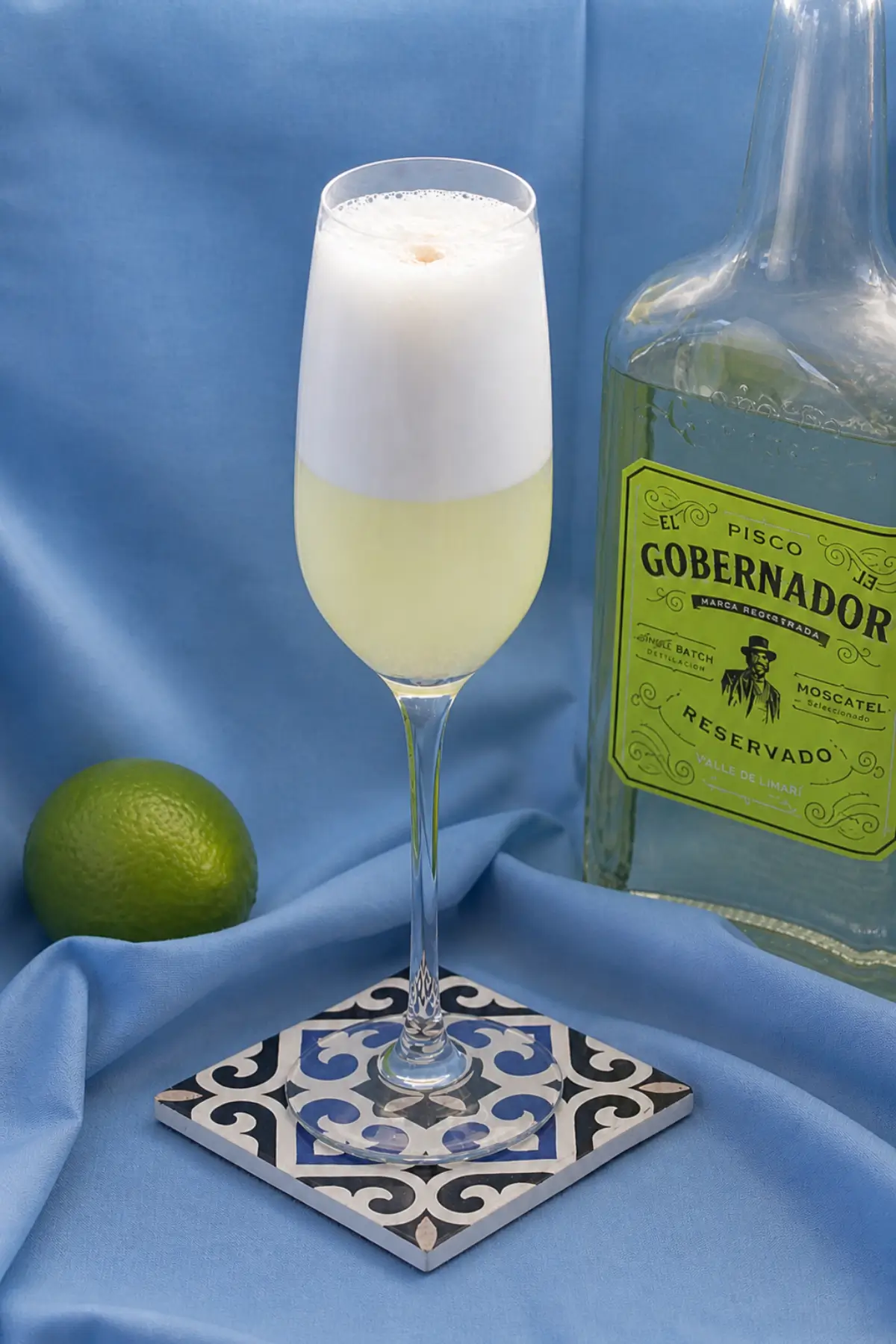 Tall glass of frothy pisco cocktail served beside a bottle of pisco and whole lime.