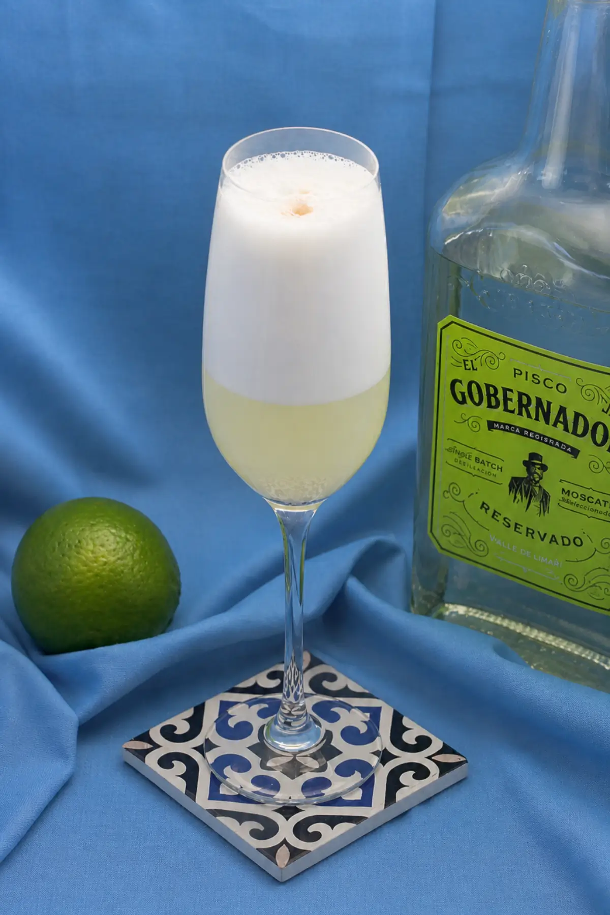 Glass of Pisco Sour with thick creamy foam served beside a bottle of pisco and fresh lime.