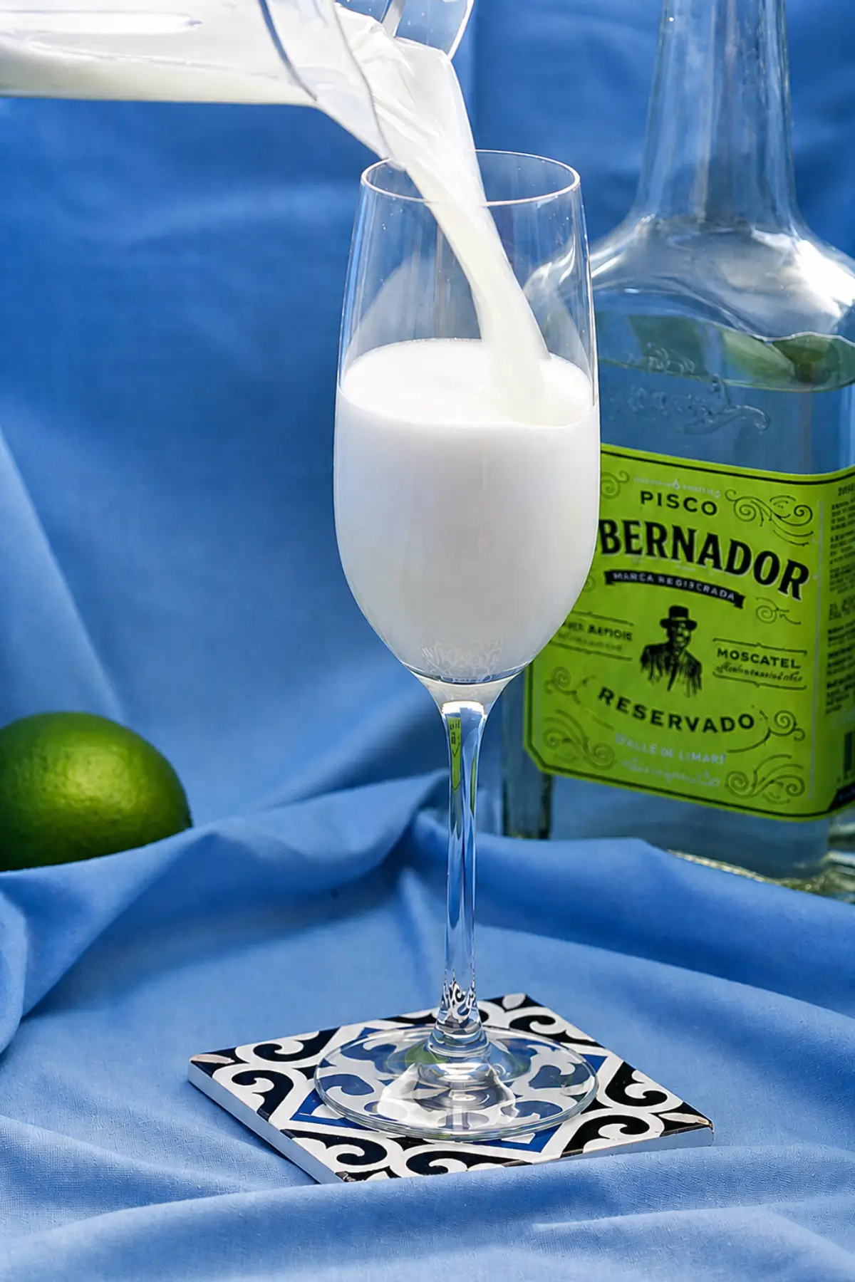 Frothy cocktail being poured into a tall serving glass beside a bottle of pisco.