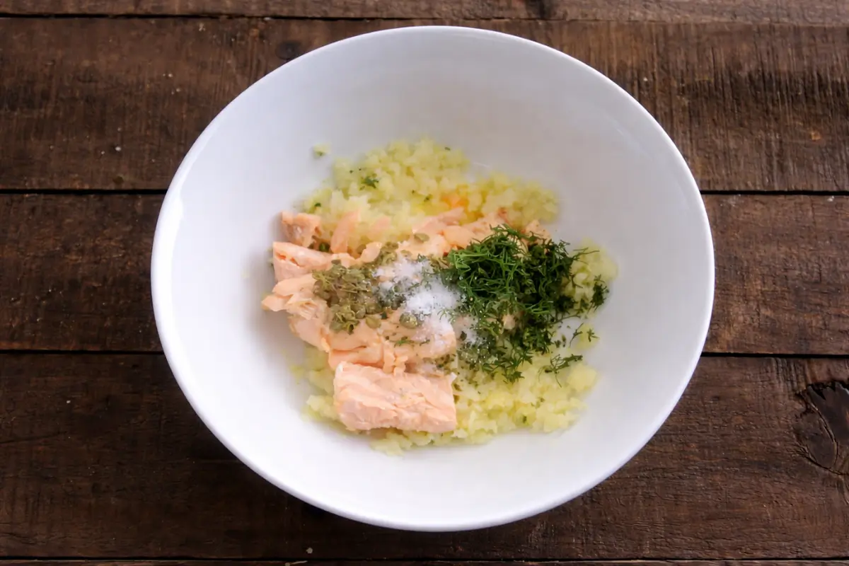 Cooked salmon mixed with mashed potato, dill, and capers in a bowl before shaping.