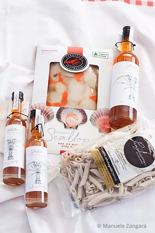 Pack of Ferguson Australia roe-on scallops with bottles of lobster-infused olive oil and wholegrain pasta on a white cloth.