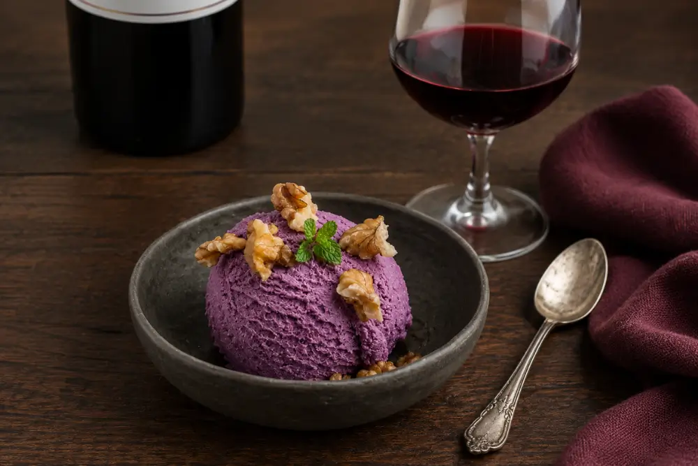Scoop of purple wine ice cream garnished with walnuts and fresh mint beside a glass of red wine.