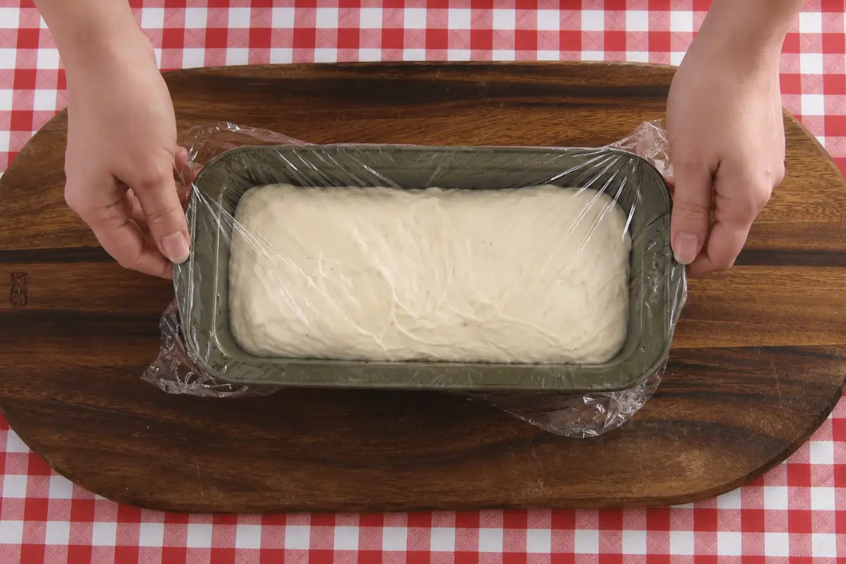 Sandwich bread dough rising in a loaf pan covered with cling wrap before baking.