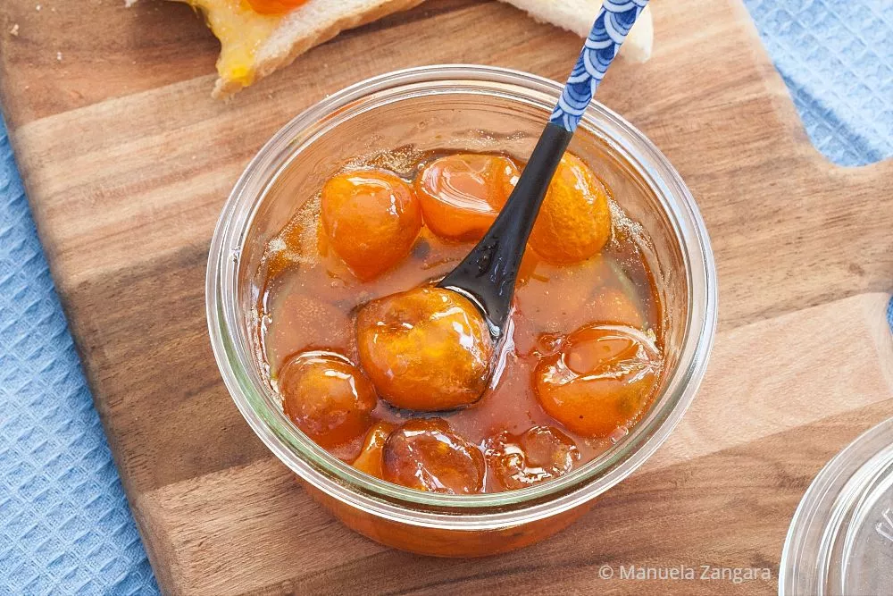Close-up of glossy kumquat jam in a glass jar with a spoon on a wooden board.