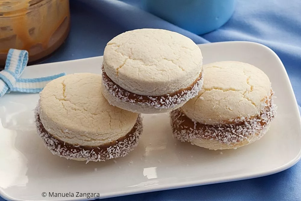 Three Alfajores de Maicena filled with Dulce de Leche and rolled in coconut, served on a white plate.