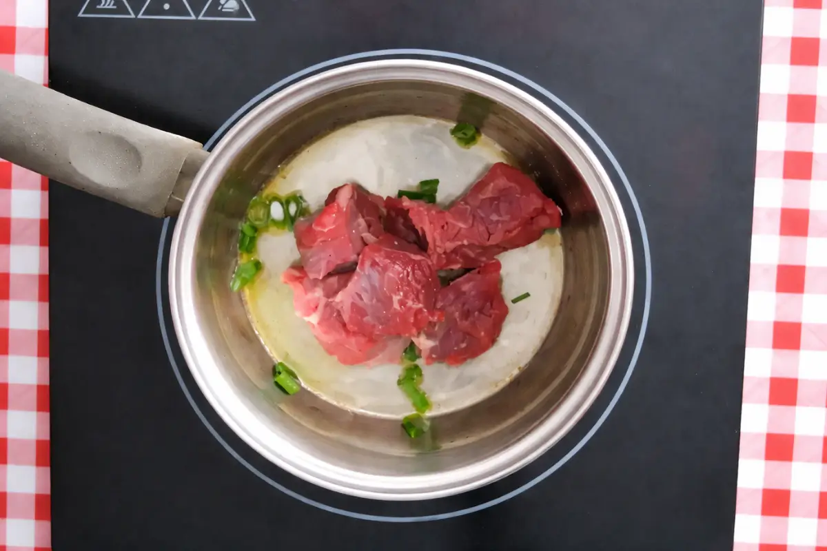 Cubed beef added to a pot with sautéed green onion tops and oil.