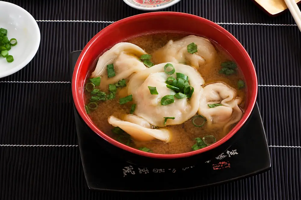 Red bowl filled with pork wontons in a light savoury broth topped with sliced green onion.