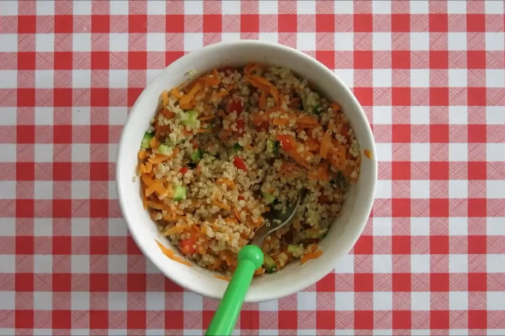 Cooked quinoa mixed with grated carrot, cucumber and capsicum in a white bowl.