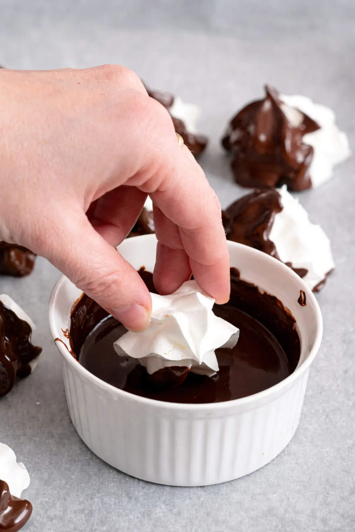 Hand dipping a baked meringue into melted chocolate on a parchment-lined tray.
