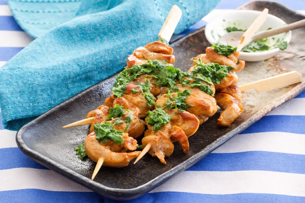 Grilled chicken skewers served on a platter with spooned herb sauce on top.