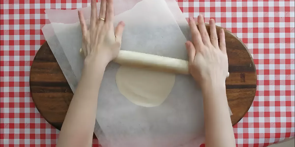 Rolling out tortilla dough between sheets of baking paper on a red-checked tablecloth.