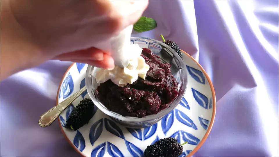 Hand piping whipped cream over a bowl of Mulberry Granita garnished with fresh mulberries and mint on a plate.
