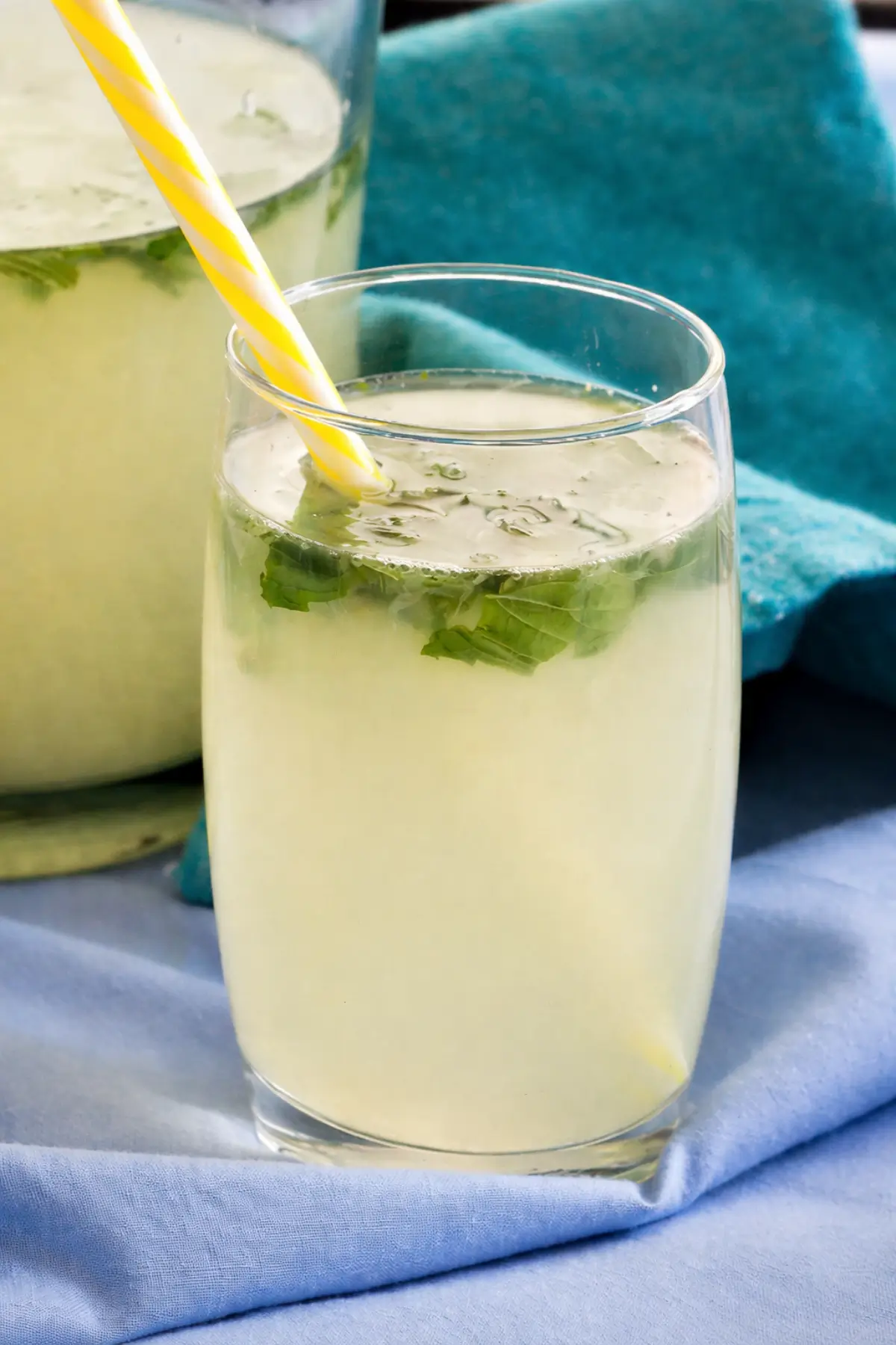 Glass of mint lemonade with mint leaves beside a filled pitcher on a blue cloth.